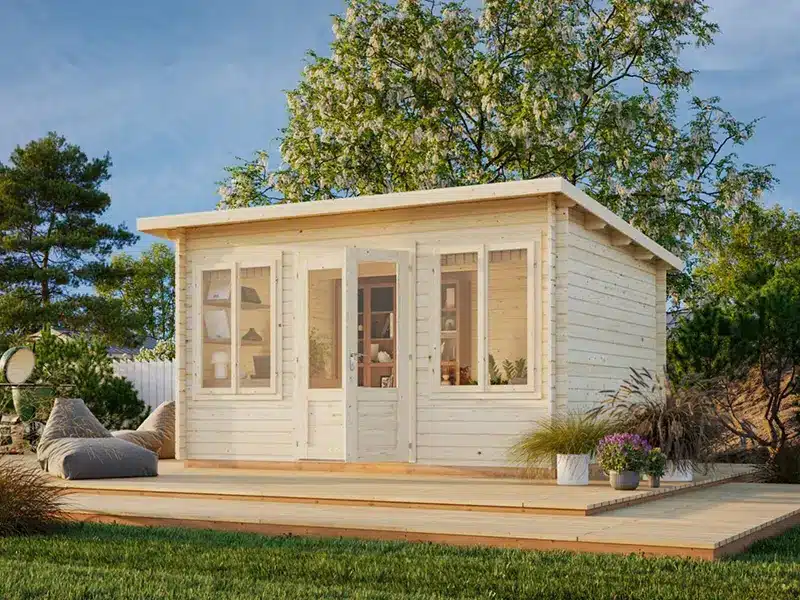 Abris de jardin en bois blanc sur terrasse en bois, avec portes et fenêtres vitrées, entouré de verdure.