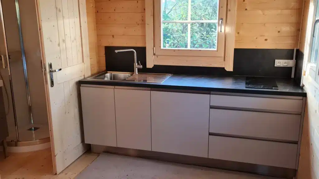 Cuisine intérieure dans une cabane en bois avec plan de travail noir et meubles gris sous une fenêtre.