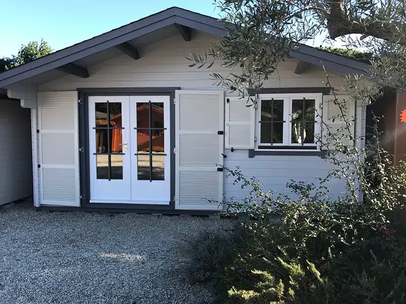 Petit chalet de jardin en bois peint blanc avec portes vitrées et volets, installé sur une allée gravillonnée près d’un arbre.