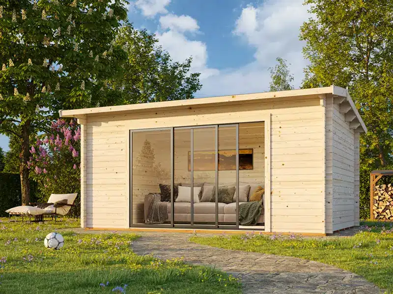 Abris de jardin en bois avec de grandes portes vitrées coulissantes et un espace salon visible à l’intérieur, posé sur une pelouse entourée d’arbres.