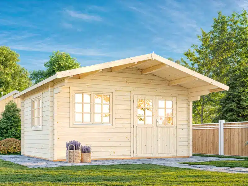 Petit chalet en bois clair avec toit en pente, porte double et fenêtres, installé sur une terrasse pavée dans un jardin entouré d’arbres et d’une clôture en bois.