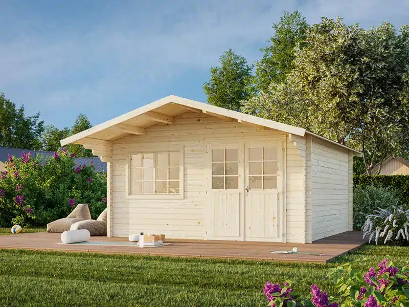 Cabane de jardin en bois blanche avec porte et fenêtres vitrées, posée sur une terrasse en bois et entourée d’herbe et d’arbres.