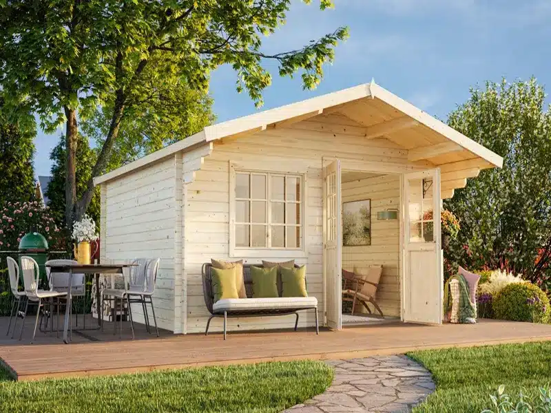 Abris de jardin en bois blanc avec terrasse couverte et canapé, sur une terrasse en bois entourée d’arbres.