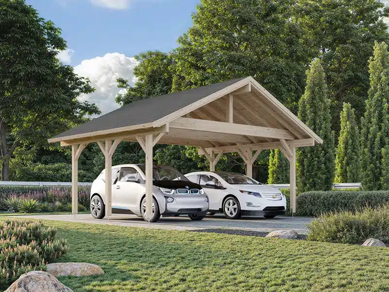 Carport en bois à toit en pente abritant deux voitures blanches dans un jardin arboré.