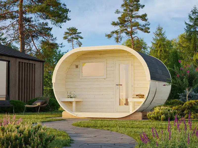 Cabine de sauna ovale en bois installée dans un jardin, avec banquette et rangements à l’intérieur.