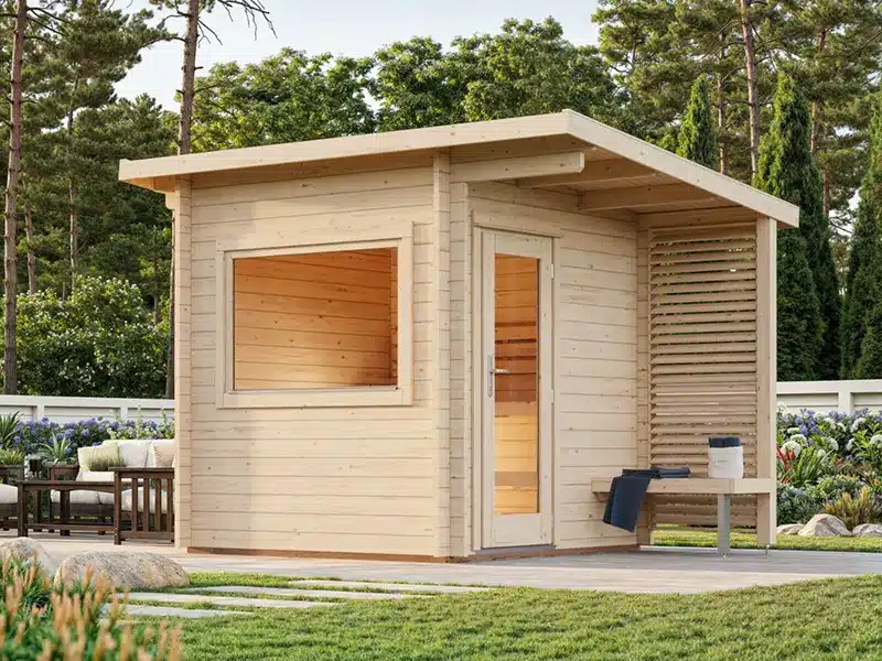 Petit abri de jardin en bois clair avec fenêtre à gauche et porte vitrée centrale, sur une terrasse partiellement couverte avec un banc, entouré d'arbres.