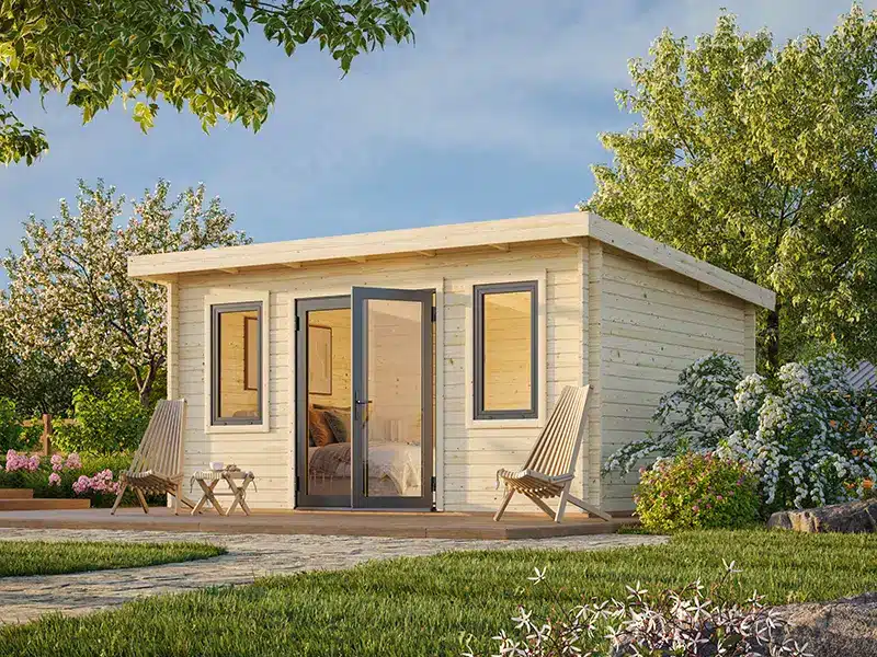 Petit chalet de jardin en bois avec porte-fenêtre et terrasse, entouré d’arbres et d’une pelouse.