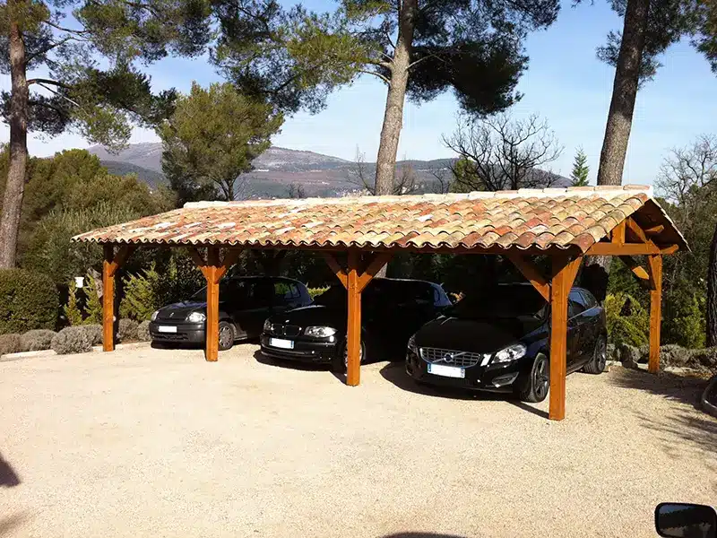 Carport en bois à toit en tuiles abritant trois voitures, entouré d’arbres et d’un paysage vallonné.