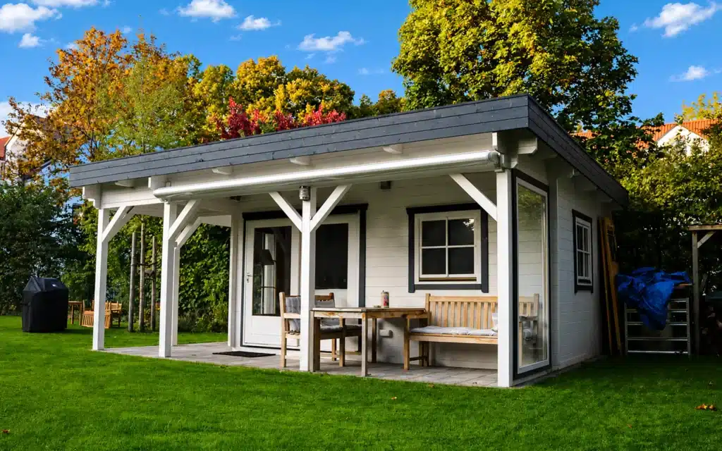 Petit chalet de jardin en bois avec terrasse couverte et mobilier, posé sur une pelouse verdoyante.