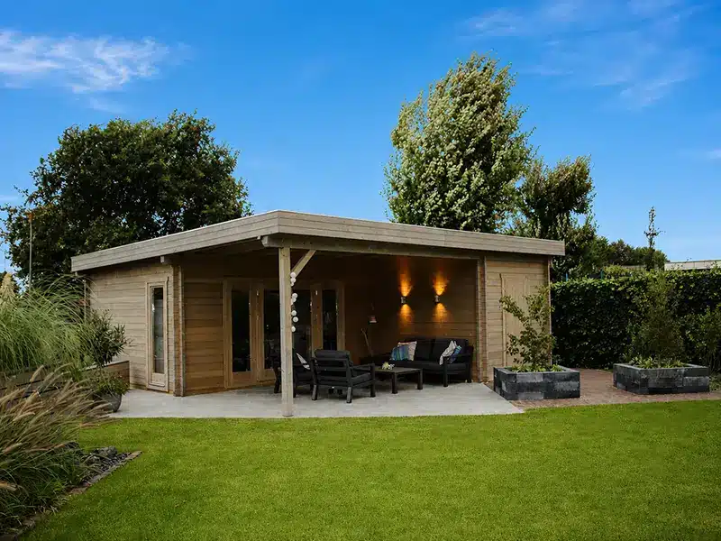 Abri de jardin en bois avec terrasse couverte et mobilier de salon, sur une pelouse verte entourée d’arbres sous un ciel bleu.